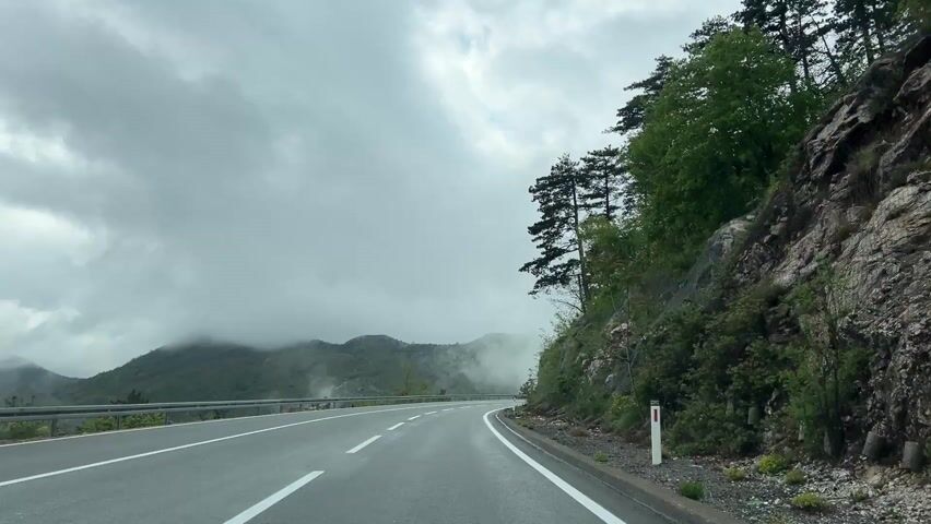 The video captures a first-person perspective from a vehicle driving along a winding mountain road. The asphalt road, marked with white dashed and solid lines, curves gently to the right. A metal guardrail runs along the left side of the road, separating it from a steep drop-off and a valley with distant, mist-shrouded mountains. To the right, a rugged, rocky hillside rises sharply, covered in green vegetation and scattered pine trees. Safety netting is visible on parts of the rock face, indicating efforts to prevent rockfalls. Small white and orange marker posts line the edge of the road on the right.

The weather is overcast, with thick grey clouds dominating the sky, and low-hanging mist partially obscuring the mountain peaks in the distance. The road surface appears damp, suggesting recent rain. As the camera progresses, two cars briefly come into view, driving in the opposite direction. The scene conveys a sense of journey through a dramatic, natural landscape under cloudy conditions. The location context is Dobrsko selo, Montenegro. There is no visible text in the scene itself.