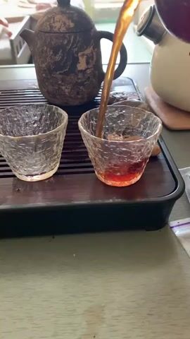 The video shows a close-up of two small glass cups being filled with dark amber liquid from a teapot. The teapot is dark and textured, sitting on a wooden tea tray. The liquid is being poured from above, with a thin stream entering the cups, causing ripples on the surface. One cup on the left is partially filled, while the cup on the right is nearly full, with the liquid reaching the rim. In the background, a white teapot or kettle is visible, along with what appears to be furniture. The surface below the tea tray is a muted green color, possibly a tablecloth or table. There are some drops of liquid spilled on the tray.