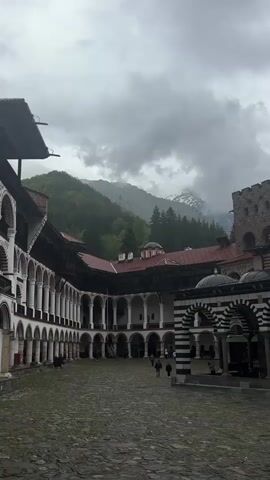 The camera captures a vertical, static shot of the interior courtyard of a monastery on an overcast day. The courtyard, paved with rough stone, is surrounded by buildings with arched colonnades. The colonnades feature white columns supporting dark eaves. Red-tiled roofs are visible on the buildings. A prominent black and white striped structure stands on the right side of the courtyard. People, identifiable by their small size relative to the architecture, walk across the courtyard. Beyond the monastery, forested mountains rise into the cloudy sky, with patches of snow visible on their peaks. The overall scene conveys a sense of history and architectural grandeur within a natural setting.