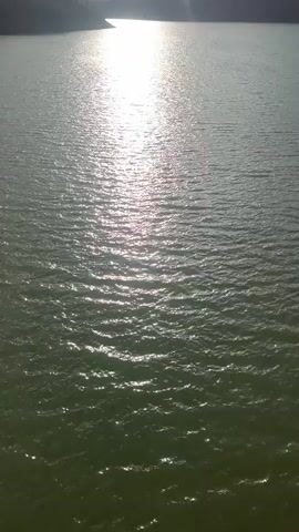 The video shows a wide body of water, likely a lake or reservoir, under a sky with scattered clouds and bright sunshine. The sun reflects intensely off the water surface, creating a shimmering effect. The surrounding landscape appears to be hilly or mountainous, with some vegetation visible along the shore. The water is calm, with small ripples visible on its surface.