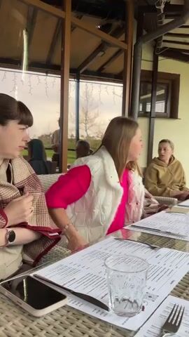 Here's a descriptive breakdown of the video:

Setting: The video is shot on the outdoor patio of a restaurant. A long table, set for a group, is the focal point. The patio is covered, with hanging string lights visible. Beyond the patio railing is a landscaped area with trees.

People: Several women are present. Some are seated at the table, looking at menus. One woman stands near the railing, looking out. Another woman walks into view briefly. They are dressed in casual clothing appropriate for a cool day.

Objects: The table is set with menus, water glasses, silverware, and a large vase filled with purple flowers. A smartphone is visible on the table. There are woven rattan chairs around the table. A backpack and other personal items are near the railing.

Actions: The women are looking at menus, apparently getting ready to order. The standing woman is observing the scenery outside.

Interactions: There appears to be conversation among the women at the table.