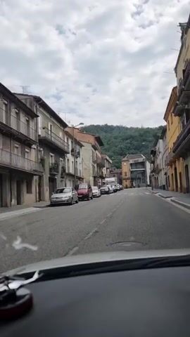 A person is driving a car down a street in a town. The street is lined with buildings on both sides, and there are cars parked along the street. In the distance, there is a hill covered in trees. The sky is cloudy.