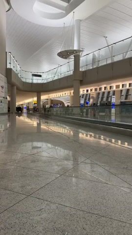 The video shows a wide shot of an airport terminal. A moving walkway is visible in the foreground, with people walking on it and beside it. The terminal has a high, modern ceiling with overhead lighting and decorative elements. In the background, shops and signs are visible, indicating a commercial area within the airport. The floor is tiled and reflective, showing the overhead lights. People are casually moving through the space, suggesting normal airport operations. The overall atmosphere is clean and spacious, typical of an international airport.