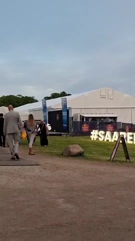 A large white tent structure with logos is visible. In front of the tent, illuminated letters spell out "#SAAREMAA". Several people are gathered in front of the tent, some are standing and talking in small groups, others are walking. The ground is a mix of dirt and grass. A large rock is in the foreground. The sky is cloudy.