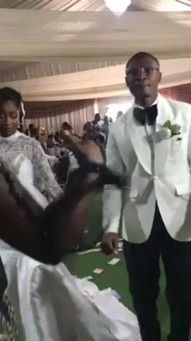 The video shows a groom dressed in a white suit and bow tie, standing on a green carpet littered with currency, at what appears to be a wedding reception inside a tented venue. A bride in a white gown stands to his left, mostly out of frame. A crowd of guests surrounds them, some dressed in traditional attire. The groom gestures with his hands as he speaks or sings along to music playing in the background. The camera moves around, focusing on different people and capturing the celebratory atmosphere of the event. The tent ceiling is draped with white fabric and a chandelier hangs above.