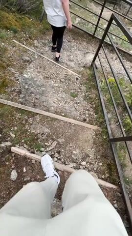 The video shows a first-person perspective of someone walking down a set of stairs in a mountainous environment. The person is wearing light-colored pants and white shoes. Another person, wearing a white shirt and black pants, is ahead on the stairs. The stairs are constructed from rocks and wooden planks, with metal railings on either side. The view from the top of the stairs reveals a vast landscape of rolling green hills and valleys under a cloudy sky. The camera pans up to capture the full scope of the mountainous view and the cloudy sky.