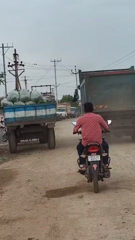 The video shows a dusty road with several vehicles, including two large trucks, a motorcycle with a rider, and some other vehicles in the background. The truck on the left is carrying a load of green produce. The truck on the right is a dump truck. The motorcycle rider is wearing a pink shirt. The sky is overcast.