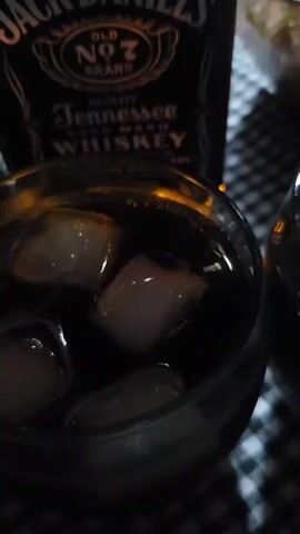 A close-up shot focuses on a bottle of Jack Daniel's whiskey and two glasses filled with a dark beverage and ice. In the background, a bowl of food and flatbread are visible on a checkered tablecloth. The lighting is dim, creating a shadowy ambiance.