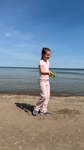 The video shows a young girl on a sandy beach. She is wearing a light pink matching top and pants, and dark sneakers. Her brown hair is braided. She is holding a green plastic bubble wand. She dips the wand into a solution and is attempting to create bubbles. The sea is calm and stretches to the horizon under a clear blue sky. The beach is mostly empty, and the girl's shadow is visible on the sand beside her.