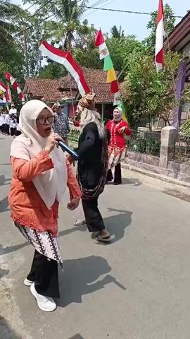 A group of people are participating in an outdoor event. In the foreground, a woman wearing a light pink hijab and an orange top with a patterned skirt performs into a microphone. To her right, a man with a long beard and a traditional headdress dances while wearing a black outfit. Another woman in a red top and similar patterned skirt is also dancing further back. The scene is decorated with Indonesian flags. The environment appears to be a street lined with houses and trees, suggesting a village or a suburban setting. The event seems to be festive, possibly a celebration or cultural performance.