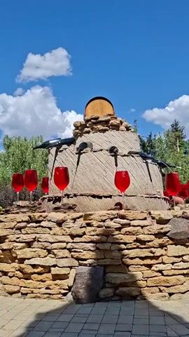 This video shows an outdoor scene on a sunny day with a blue sky and white clouds. The central focus is a large, circular, stone structure that resembles a millstone, topped with a wooden barrel. In front of the stone structure, there is a tiered rock formation. Scattered around the rock formation and the base of the millstone are several oversized, red wine glasses. The overall impression is a decorative landscape feature, possibly in a garden or winery.