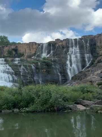The video pans across a wide, multi-tiered waterfall cascading down a rocky cliff face. The water is white and foamy, creating a powerful visual effect. Lush green vegetation, including trees and bushes, surrounds the base of the waterfall and grows on the cliff itself. In the foreground, the calm surface of a body of water, possibly a river or lake, reflects the scene. The sky above is partly cloudy with patches of blue. There are no people visible in the video. The scene appears to be a natural landscape.