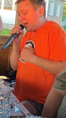 A man in an orange t-shirt is holding a microphone to his mouth and singing. To his left, another man in a patterned t-shirt and a hat is pouring a beverage from a green bottle into a glass. The beverage appears to be foamy. The scene appears to be outdoors, under a wooden canopy, with a building visible in the background. The lighting suggests it is late afternoon or early evening. There are cans and glasses on a table in the foreground.