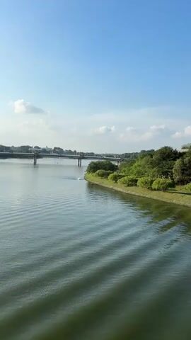 The video captures a serene view of a wide, calm body of water with gentle ripples, moving from left to right. On the right bank, a lush green park with manicured trees and grass slopes down towards the water. Further inland, a grand, colonial-style building with a distinctive green dome sits atop a hill, surrounded by dense foliage. In the distance, a bridge spans the water, with faint structures visible on the far bank. The sky above is a clear, vibrant blue with scattered white clouds. No people or specific actions are clearly discernible.