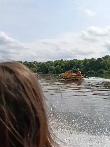 The video shows a person from behind, their brown hair obscuring much of the view, looking out at a body of water. They appear to be on a boat, indicated by the churning wake behind them and the edge of the boat visible in the foreground. In the distance, another boat, which is orange and white, is visible on the water. The background consists of a tree-lined shore under a cloudy sky. The camera is focused on the water and the distant boat, with the person in the foreground serving as a framing element.