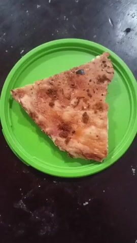 A single slice of pizza sits on a bright green plastic lid. The pizza appears to be a thin crust with a reddish sauce and sparse toppings, some of which are dark brown and crumbly. A small dark object, possibly a fly or a crumb, is visible on the surface of the pizza. The lid is circular and has a raised rim. The background is a dark, textured surface, likely a table. The lighting suggests an indoor environment. The video is a static shot, with no visible movement.
