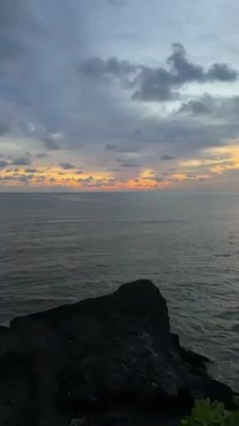 The video shows an ocean horizon with clouds lit by the setting sun. In the foreground, there is a dark, rocky cliff and some green foliage in the lower right corner of the frame. The sky is cloudy, and the clouds are dark with streaks of orange and yellow where the sun is setting on the horizon. The ocean water is gray, and some ripples are visible on the surface. The camera is static, providing a stable view of the scene.