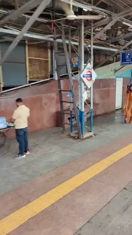 A train station platform is shown. A digital clock displays 12:44. Several people, mostly women, are walking along the platform, carrying bundles on their heads and shoulders. Some are wearing traditional Indian clothing. A man in a green shirt walks past with a phone in his hand. A train car is visible on the right side of the frame. The platform has a yellow tactile warning strip along the edge. Murals are painted on the walls behind the passengers.