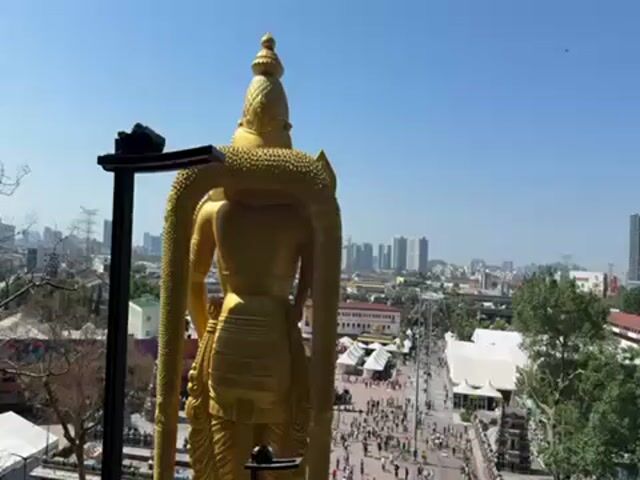 The video provides an elevated view, taken from above, overlooking a crowd of people walking along a path and ascending stairs. A large, golden statue of a deity stands in the foreground, its back to the camera, appearing to guard the scene below. Beyond the crowd and path, a city skyline with several high-rise buildings is visible under a clear, blue sky. Green foliage is present on the right side of the frame, adding a natural element to the urban and religious setting. The overall atmosphere is one of a busy, possibly touristy, location on a bright, sunny day.