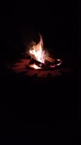 A campfire is burning in a circular fire pit made of bricks. The fire is bright and lively, with flames reaching upwards and smoke billowing around them. The surrounding area is dark, indicating that it is nighttime. The scene is peaceful and tranquil.