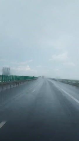 The video shows a first-person perspective of driving on a highway in rainy weather. The road is wet and reflective, with streaks of water visible. The sky is overcast and gray. Guardrails are visible on the left side of the road, and there are other vehicles in the distance. The windshield appears to have raindrops on it, causing some blurriness. The overall atmosphere is somber and muted due to the weather conditions.
