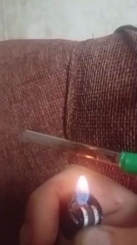 The video shows a close-up view of a person's hand holding a lighter that is ignited. The flame of the lighter is blue and white, and it is being held near a glass pipe. The pipe contains a substance that is being heated and appears to be vaporizing. The background of the video is a brown, textured fabric, possibly upholstery. The lighting is dim, creating a focused view on the hand, lighter, and pipe.