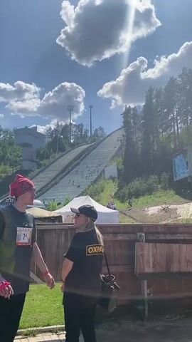 The video shows a ski jump on a sunny day with scattered clouds.  Several people are visible on the slope.  Two people are standing in the foreground; one is wearing a black shirt and a red bandana, and the other is wearing a black cap and a security shirt with the word "OXPAHA" written in yellow.  A white tent and wooden fence are also in the foreground.  Tall trees flank the ski jump.  There are spotlights atop the jump and a digital display board on the right.