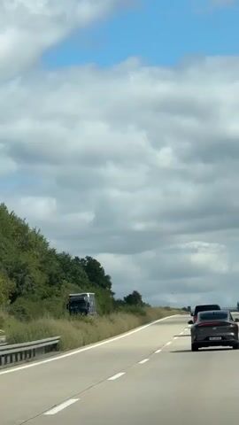 A dark colored sedan is driving on a highway. The vehicle is in the right lane of a two-lane highway. The car is following another vehicle that is not fully visible. The road is paved with asphalt and has white lane markings. The highway is bordered by a grassy median on the left and trees and vegetation on the right. The sky is blue with scattered clouds. The weather appears to be sunny. There are other vehicles visible in the distance, as well as highway signs.