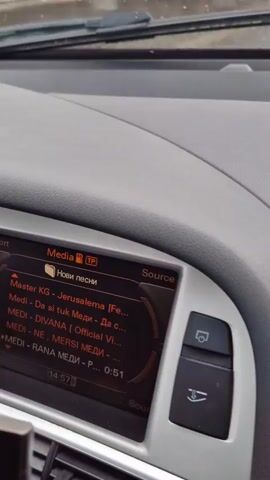 From inside a car, the screen of a car infotainment system is shown playing music. The screen displays a folder called "New songs" and lists several songs by the artist "MEDI" and one by "Master KG." The wipers are visible at the top of the screen, suggesting it is raining. Part of a smartphone in a holder is visible in the lower left of the frame.