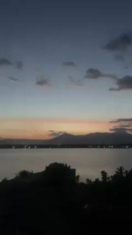 The video shows a crescent moon in the sky. The sky is a gradient of blue at the top, fading to a pinkish-orange near the horizon. Below the sky, a body of water is visible, reflecting some of the light. In the distance, there are dark land masses, possibly mountains, silhouetted against the horizon. The foreground shows some silhouetted trees and greenery. The scene appears to be taken during dusk or dawn.