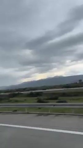 The video shows a view from a moving vehicle, likely a car, on a highway. The scenery consists of a wide, meandering river and a vast green field with scattered trees, extending towards a distant mountain range under a cloudy, overcast sky. The gray metal highway barrier runs parallel to the road, and the gray asphalt surface of the highway includes painted white lines.