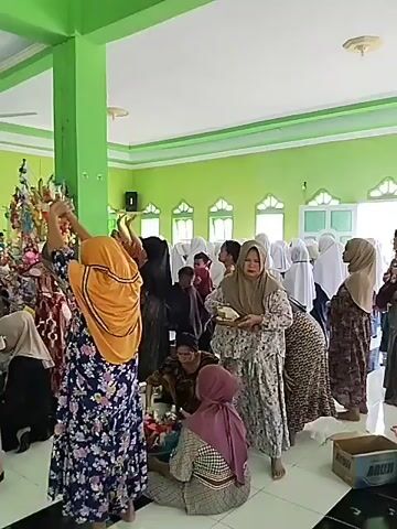 A group of people, predominantly women, are gathered in a brightly lit room with green walls. Some individuals are wearing headscarves and traditional Islamic attire. In the foreground, a woman in a patterned dress and headscarf is holding a plate of food. To the left, a woman in a yellow headscarf and a floral dress is bent over. Several decorated trees or displays, adorned with colorful items and what appear to be papers or small objects, are prominent in the center of the room. In the background, more people, including some men in white clothing and skullcaps, are visible. The room appears to be a community hall or a place of worship, possibly a mosque, with ceiling fans and decorative elements on the walls. Some people are sitting on the floor, while others are standing. The overall atmosphere suggests a communal gathering or event.
