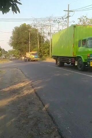 The video shows a rural road with a few vehicles. On the right side of the road, a large green shipping container is being transported by a truck. In the center of the road, a yellow and blue truck is driving with a person on a motorcycle behind it. Several other motorcycles are also present on the road, some of which appear to be following the truck. The road is paved and has some damage. Trees and utility poles are visible on the sides of the road. The sky is clear and the sun is shining.