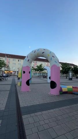 The video shows a colorful, inflatable playground structure resembling a giant worm or pretzel with a frosting-like topping and sprinkles. The structure is installed in an outdoor public space with paved ground and buildings in the background. The sky is a pale blue, suggesting dusk or early morning. The structure has two archways with holes for children to pass through, and a segmented, colorful body extending along the ground. The faces on the archways appear to be cartoonish, with wide-open mouths. The video pans from left to right, showing different angles of the structure and its surroundings. No people are visible in the immediate vicinity of the playground. The overall scene is calm and quiet.