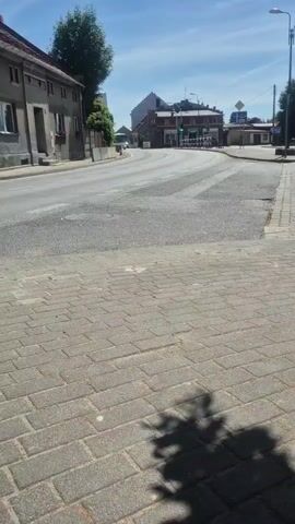 This is a horizontal, handheld video recording filmed from a sidewalk. The camera is angled slightly downwards towards a cobblestone sidewalk, with a shadow of a tree cast on it. Across the sidewalk is a paved road with cars, and on the other side of the road are buildings, including a multi-story building with a red-tiled roof and a large tree. Further down the road, a bus is visible, approaching an intersection with traffic lights. The sky is blue with a few scattered clouds, suggesting a clear, sunny day. The overall setting appears to be a small town or village, possibly in Bralin, Poland, given the context. The scene is peaceful and ordinary, depicting a typical daytime street view. There is no discernible text visible in the video.