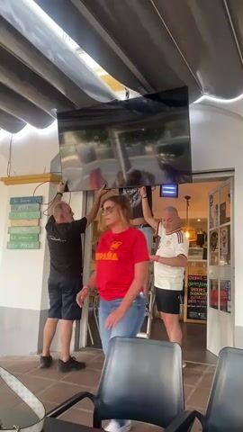 This video shows a group of people installing a large flat-screen television on a ceiling mount. Two men are actively holding the television in place. One man, wearing a black t-shirt and shorts, stands on a step ladder, supporting the TV from below. Another man, wearing a black t-shirt and shorts, stands to the left of the ladder, also holding the TV. A third man, wearing a white and black striped shirt and shorts, stands to the right, looking on. A woman, wearing a red t-shirt and jeans, is visible in the foreground. The scene appears to be an outdoor or semi-outdoor seating area, possibly a restaurant or bar, with a canopy overhead and a doorway visible in the background. Cables are hanging from the TV, suggesting it is being connected.