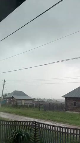This media file captures a rural scene in Urazovo, Russia. The primary subjects are two traditional wooden houses with dark metal roofs, situated amidst green grass and dirt paths. A wooden fence runs across the foreground, partially obscuring a view of the houses and the surrounding landscape. The sky is overcast with heavy grey clouds, indicating a dreary or rainy day. The overall atmosphere is somber and quiet.

The activity taking place is implied by the weather. The rain is visible as streaks falling from the sky, hitting the ground and the surfaces of the houses and fences, creating a wet sheen. The scene depicts a moment of natural weather event impacting a rural settlement.

Notable details include the rustic architecture of the wooden houses, characteristic of rural Russian villages. The presence of utility poles and power lines suggests that this is a inhabited area, albeit a sparsely populated one. The dampness and the cloudy sky contribute to a sense of isolation and perhaps melancholy. There is no visible text in the media file. The time of day is difficult to ascertain precisely due to the overcast conditions, but it appears to be daytime.