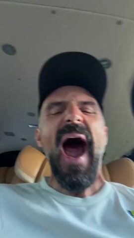The media file is a close-up, low-angle video of a man wearing a black baseball cap and a white t-shirt. He has a graying beard and mustache. The man's face is the primary subject, and he makes a series of expressions: laughing with his eyes closed, looking straight ahead with his eyes closed, looking ahead with his eyes open, widening his eyes in surprise, and finally looking ahead with a neutral expression.

The setting appears to be the interior of a vehicle, possibly a private jet or a high-end van, given the beige leather seats visible behind him and the overhead lighting fixtures. The lighting suggests it is daytime, with natural light filtering in, creating highlights on the man's face. There is no visible text in the video. The man's expressions shift from amusement to surprise and back to a neutral state, suggesting he is interacting with someone or reacting to something off-camera.