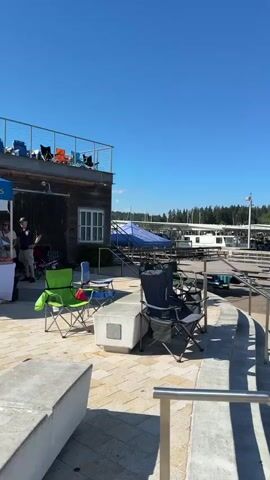 The video pans across a sunny outdoor scene at a marina on a clear day with a bright blue sky. In the foreground, there is a paved area with a curving walkway bordered by metal railings and concrete planters. Several folding chairs are set up on the paved area and a grassy slope leading down to the water. Beyond the chairs is a wooden dock with several people standing on it, and beyond the dock, a body of water with numerous boats docked and moored. Further in the background, a tree-covered hillside rises, with houses visible amongst the trees. A building with a red roof is situated on the water's edge, and a large white pole stands prominently in the middle ground. Several people are visible on the dock, and one person in a yellow hat and pink shorts is walking on the grass. Another person is standing on a paddleboard in the water. The overall impression is a lively, recreational waterfront setting.