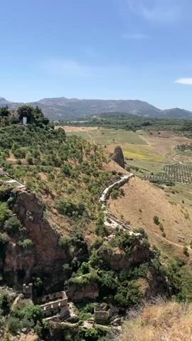 The video shows a panoramic view of a town perched on a cliffside. The town is characterized by white buildings with red-tiled roofs, typical of a Mediterranean architectural style. The buildings are situated along the edge of a steep, rocky cliff, with lush green vegetation growing on its slopes. Below the town, the landscape opens up to a valley with rolling hills, agricultural fields, and distant mountains under a clear blue sky. A winding path is visible on the side of the cliff, suggesting a way to access the town or explore the surrounding area. Some ruins of older structures are scattered on the lower parts of the cliff. The overall impression is one of a picturesque and historic settlement nestled within a dramatic natural setting.