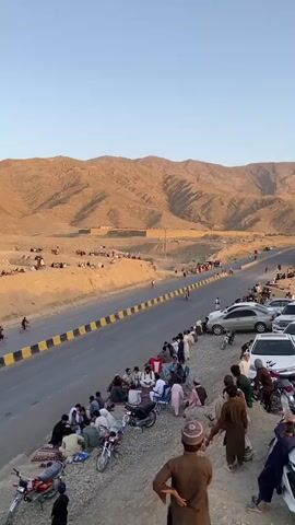 The video shows a roadside scene with a group of people gathered on a hillside overlooking a road and a desert landscape with mountains in the background. The people are mostly men, some sitting on rugs and chairs, others standing around motorcycles. The road has yellow and black barriers. On the other side of the road, more people are gathered on a sandy area, some watching cyclists on the road. Several cars are parked near the group on the hillside. The sky is clear and the light suggests it is late afternoon or early evening.