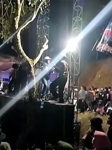 A group of people are gathered around a stage at night. Two individuals are on the stage, one in a blue jacket and the other in a black shirt. A bright spotlight shines from above, illuminating the stage and the people on it. The background shows a crowd of people, suggesting a performance or event is taking place. The environment appears to be outdoors, with trees and foliage visible. A large speaker is positioned in front of the stage.