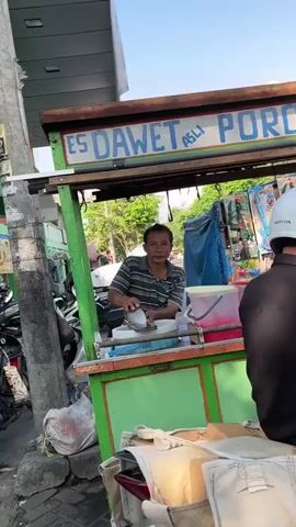 The video shows a street vendor preparing a drink. The vendor, a man with short dark hair, is standing behind a green cart with "ES DAWET ASLI PORONG" written on it. He is wearing a striped shirt and appears to be adding ingredients to a clear plastic cup. Several motorcycles are parked nearby, and there are trees and buildings in the background. The scene suggests a bustling urban environment.