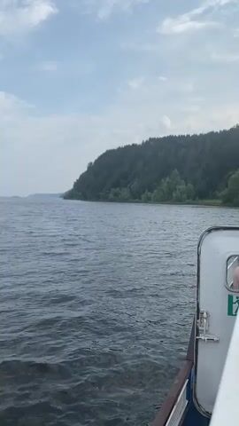 A first-person perspective video shows a view from a boat moving on a river. The water is a deep blue and slightly rippled. On the right side of the frame, the white side of the boat is visible, with a door or hatch and a person partially visible through a window. The left side of the frame shows a vast expanse of water leading towards a distant, tree-covered hill or mountain range. The sky is a pale blue with scattered white clouds. The overall impression is one of a scenic river cruise on a slightly overcast day.