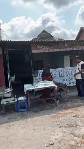 The video shows a street scene in front of a food stall or restaurant. A woman wearing a hijab and a red shirt is behind a table with food items, appearing to serve a customer. A man in a white and black shirt and dark pants stands in front of her, facing her. Behind them is a building with a sign that says "BAKSO H. M. SUKRI" in large letters, with smaller text below. The environment appears to be an outdoor area with a paved ground, possibly a street or sidewalk. There are other tables and chairs visible, suggesting a seating area for customers. The overall impression is of a busy commercial area.