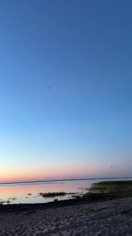 The video shows a sunset over a body of water. The sky transitions from a deep blue at the top to shades of orange and pink near the horizon. The water is calm and reflects the colors of the sky. A sandy beach is in the foreground, with some vegetation and what appear to be posts or a small structure in the distance. The video is filmed vertically and appears to be from a moving vehicle or with handheld camera movement.