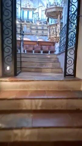 The video shows a slow ascent of a stone staircase within a church. The staircase is flanked by ornate black metal railings. At the top of the stairs, a view opens up to the interior of the church. In the background, a large pipe organ with golden embellishments is visible above a row of dark wooden chairs. To the right of the organ, a pulpit, also intricately decorated, is mounted on the wall. The church interior features high vaulted ceilings painted with frescoes and is lit by overhead lights. The floor visible at the top of the stairs has a checkered pattern.