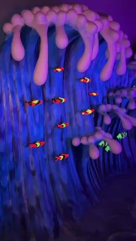 The video shows a brightly colored, blue wave with abstract white foam. Artificial fish in various colors like red, yellow, and neon green appear to swim amongst the waves. The video's lighting suggests a blacklight or similar effect, making the colors stand out. The overall environment has a playful, underwater-themed aesthetic.