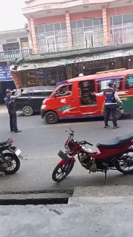 Two individuals wearing blue vests with orange markings are directing traffic on a street. One of them, on the left, stands near a red and green van, while the other, on the right, gestures with his hand towards a black SUV. Several motorcycles are parked on the side of the road. In the background, a multi-story building with storefronts lines the street. The scene appears to be a typical urban environment with ongoing vehicular and pedestrian activity.
