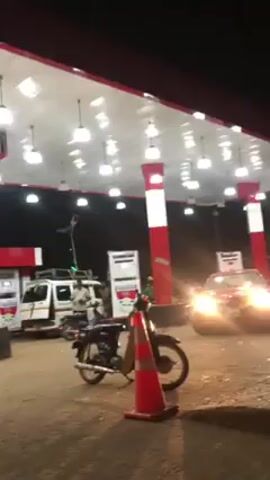 The video shows an outdoor gas station at night. Several motorcycles are parked near orange cones. A white van with items on top and a red car with its headlights on are also present. A person walks into the frame. The gas station has a red and white color scheme with multiple lights illuminating the area. The ground appears to be dusty or dirt. The video shakes.